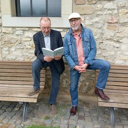 Das Bild zeigt Reinhard Försterling und Martin Krüger-Düsenberg vor einer Steinfassade, beide haben ihre Füße jeweils auf eine Bank gestellt.